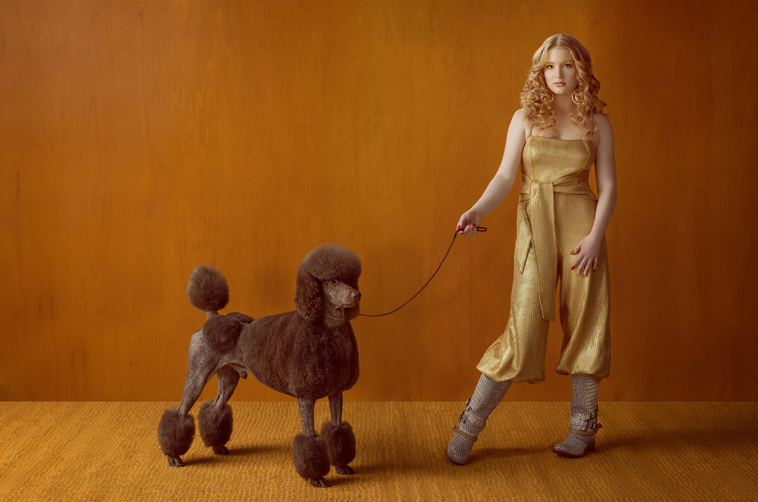 Female model standing holding a leash with a brown standard poodle. 