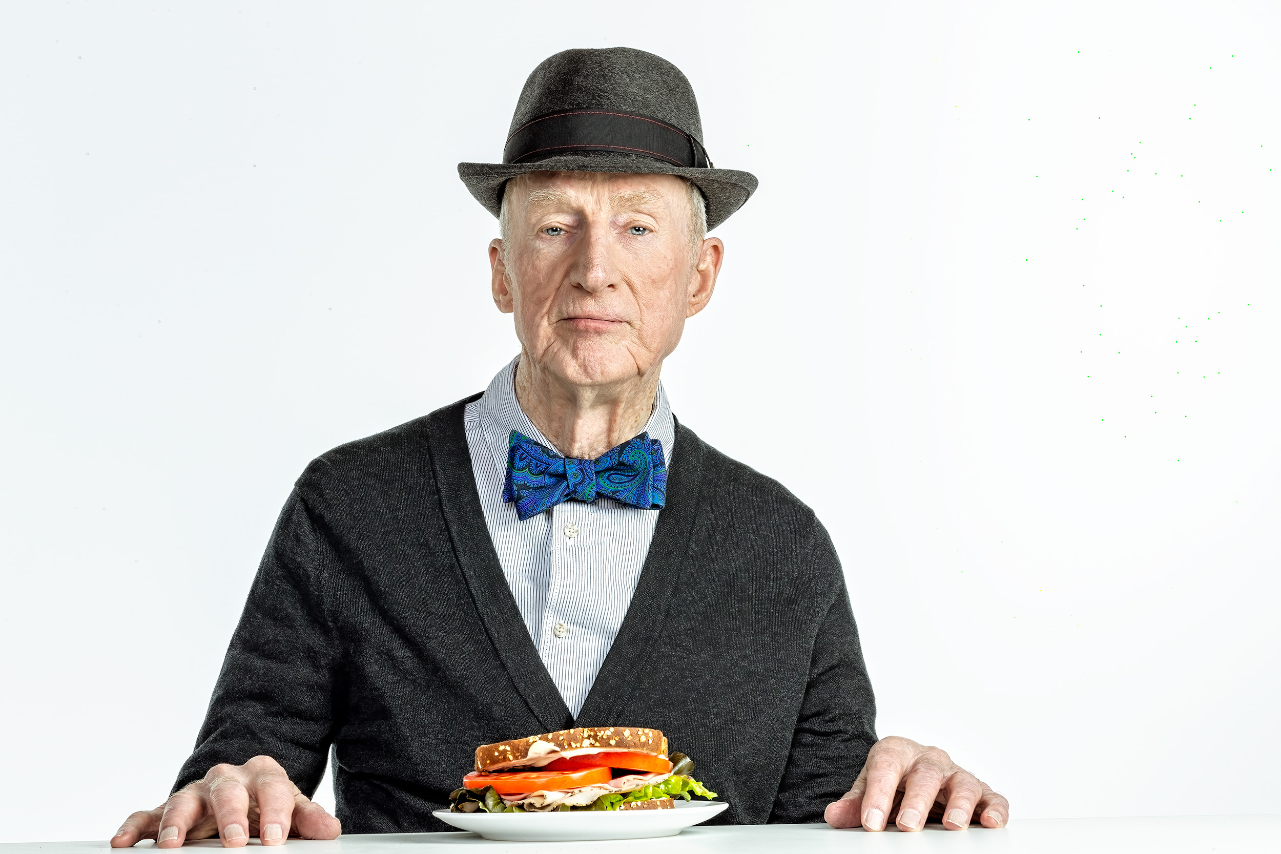 Elder man with bowtie and sweater wearing a hat at a table with a tomato sandwhich Community Table