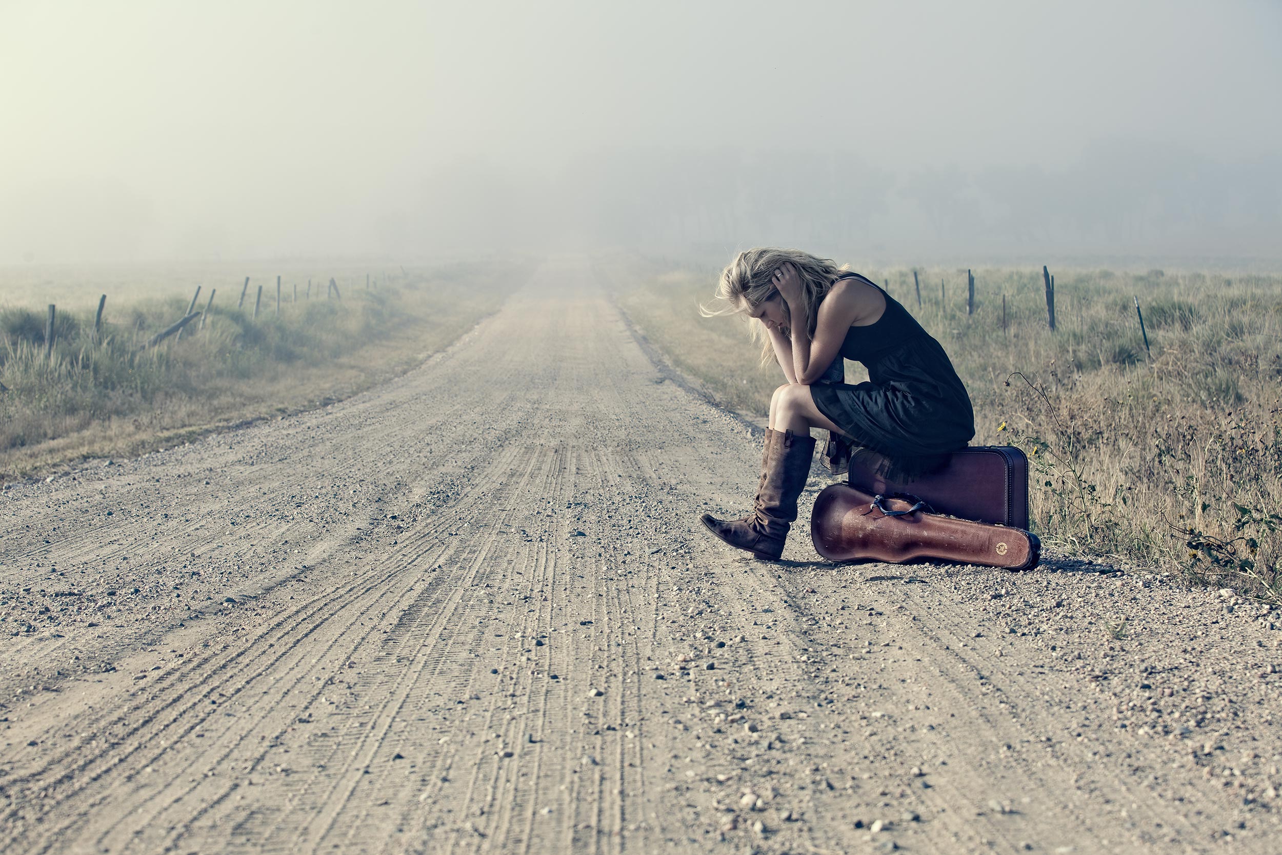 Female blonde model sitting with violin case on an empty road. 
